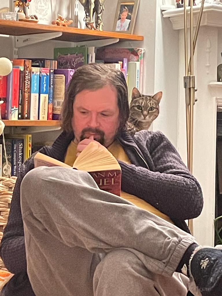 Image shows Mark Sheerin reading a book while a cat looks over his shoulder